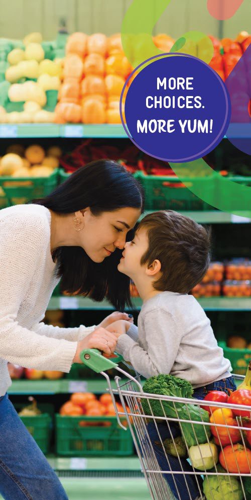 An adult and child smile at each other in a grocery store. The child sits in a shopping cart filled with fruits and vegetables, with colorful produce displayed behind them. A circular graphic reads “More choices. More yum!”