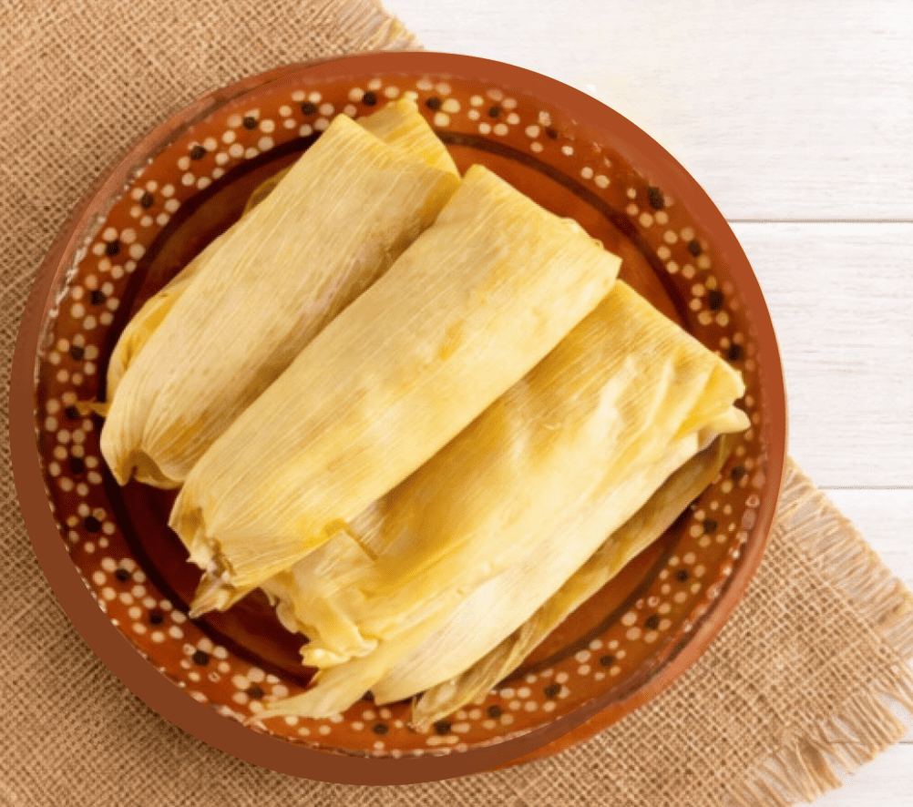 Plate of corn‑husk‑wrapped tamales on burlap over a light wooden surface.