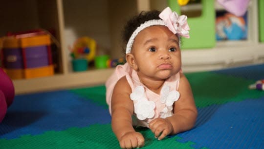 Baby lying on a colorful foam play mat, wearing a pink outfit and headband in a playroom.