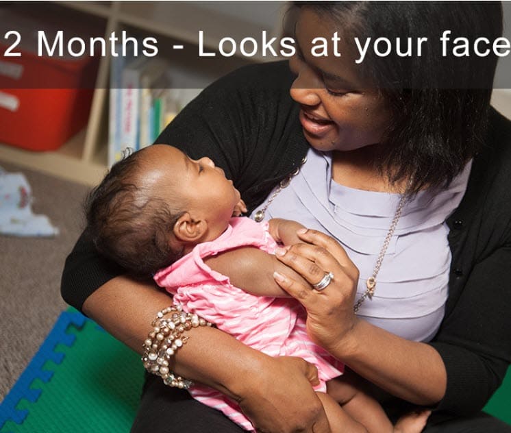 Adult smiling at a young baby who is looking up at their face in a playroom.