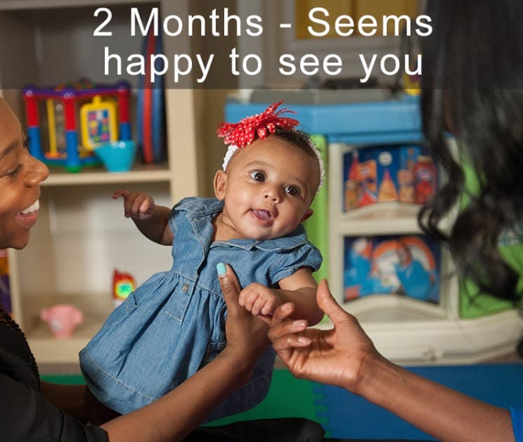 Adult holding a baby in a playroom while another adult reaches toward the smiling baby.