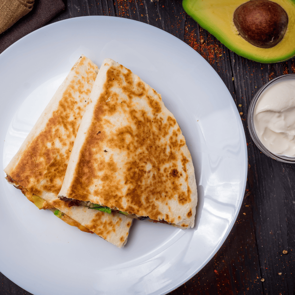 Two folded quesadillas on a white plate with an avocado half, sour cream, and seasoning on a dark wooden surface.